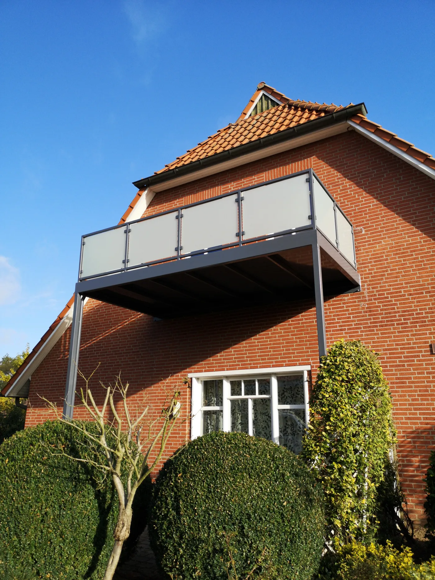 Anbaubalkon mit anthrazitfarbenem Rahmen und Milchglasfüllung vor einem Klinkergiebel, vorn auf zwei schlanken Stützen, unter strahlend blauem Himmel.