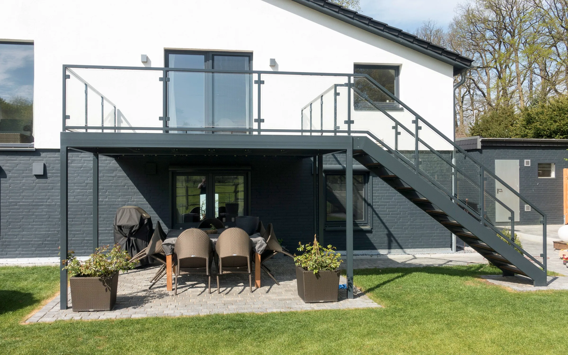 Anthrazitfarbener Anbaubalkon mit Glasgeländer und gerader Außentreppe in den Garten, darunter eine überdachte Terrasse mit Gartenmöbeln. Gebaut in Barendorf.