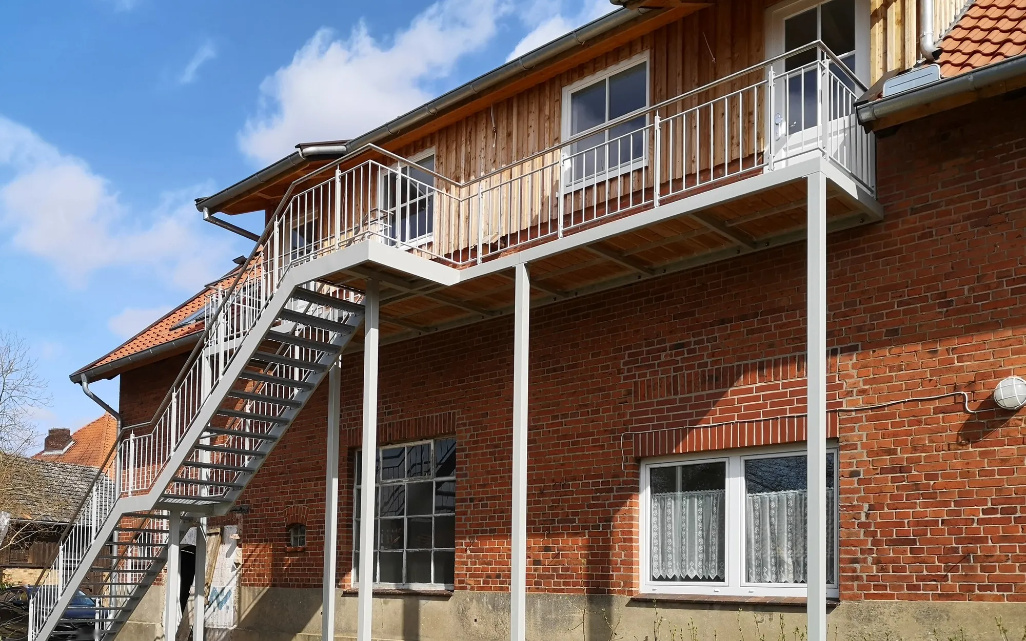 Lange Balkonanlage mit Stabgeländer auf schlanken Stützen und gerader Außentreppe an einem umgebauten Klinker-Resthof mit Holzgiebel. Gebaut in Bienenbüttel.