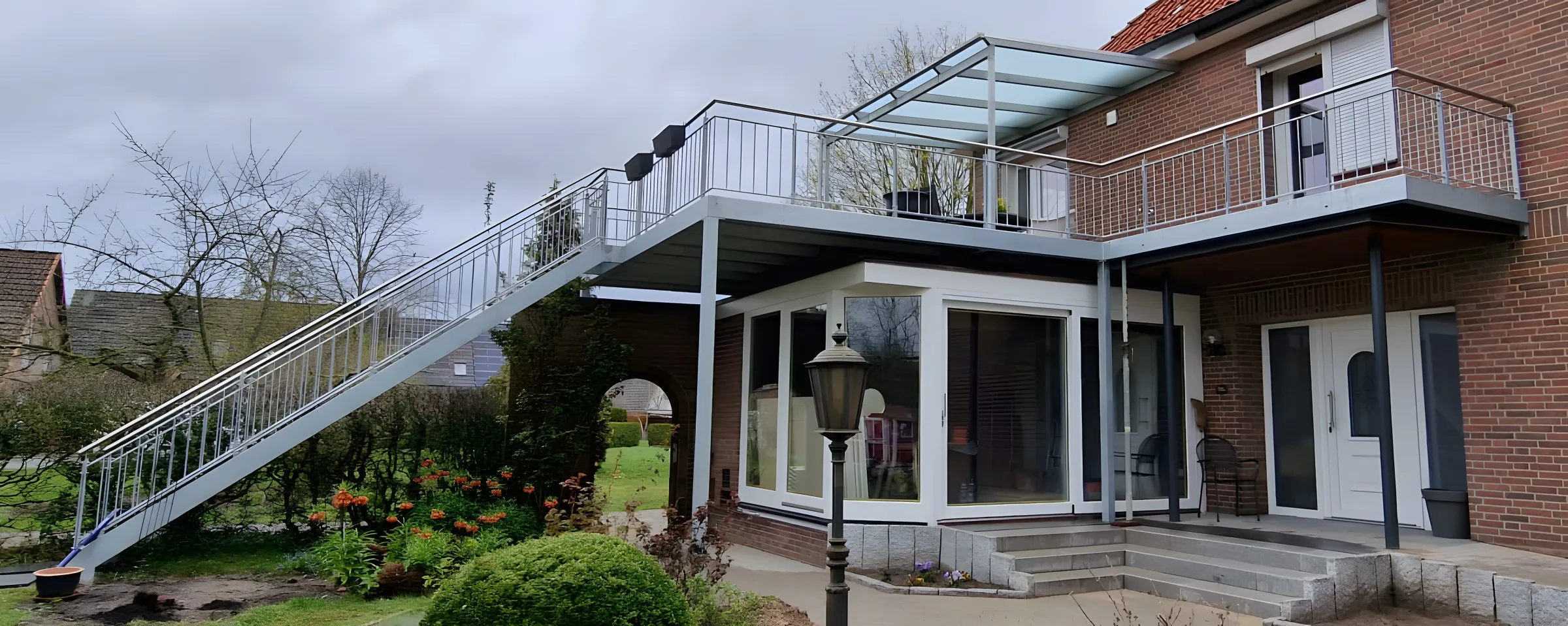 Anbaubalkon auf Stützen mit Stabgeländer, Glasüberdachung vor der Balkontür und langer Außentreppe in den Garten eines Klinkerhauses. Gebaut in Artlenburg.