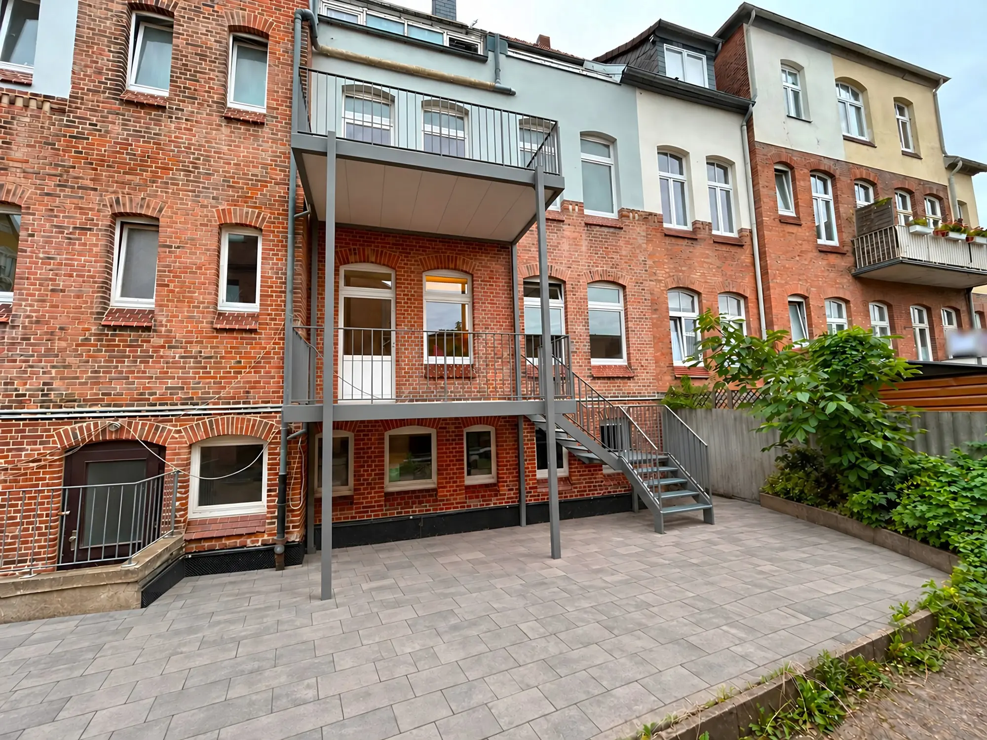 Fertiger Vorstellbalkon über zwei Geschosse an einem Klinkerhaus: zwei Balkone auf schlanken grauen Stützen mit Stabgeländer, dazu eine Außentreppe mit Podest zur gepflasterten Terrasse