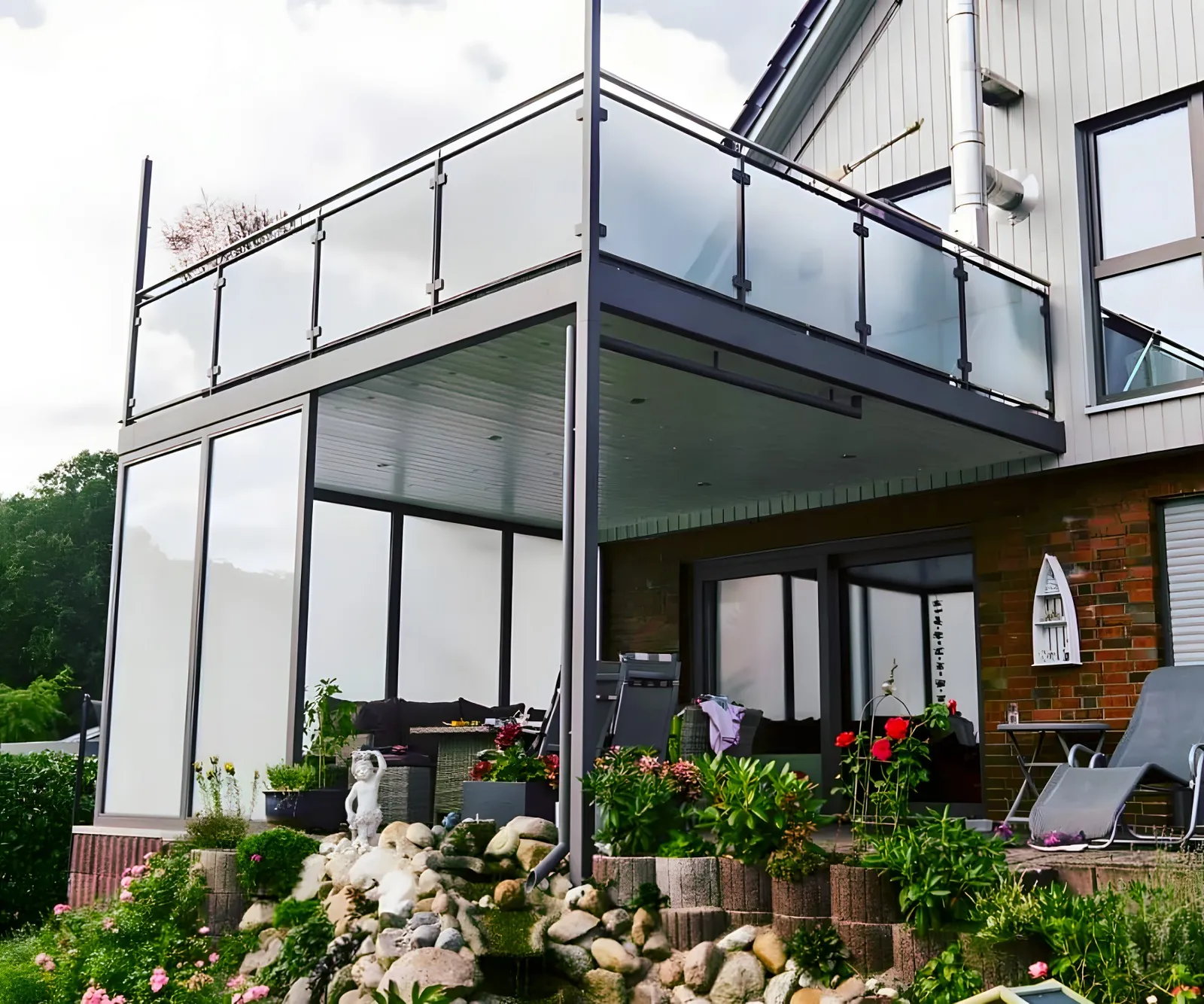 Balkon mit Glasgeländer an einem Haus mit Holzfassade, darunter eine rundum verglaste Terrasse, vorn ein Steingarten. Gebaut in Lüneburg.