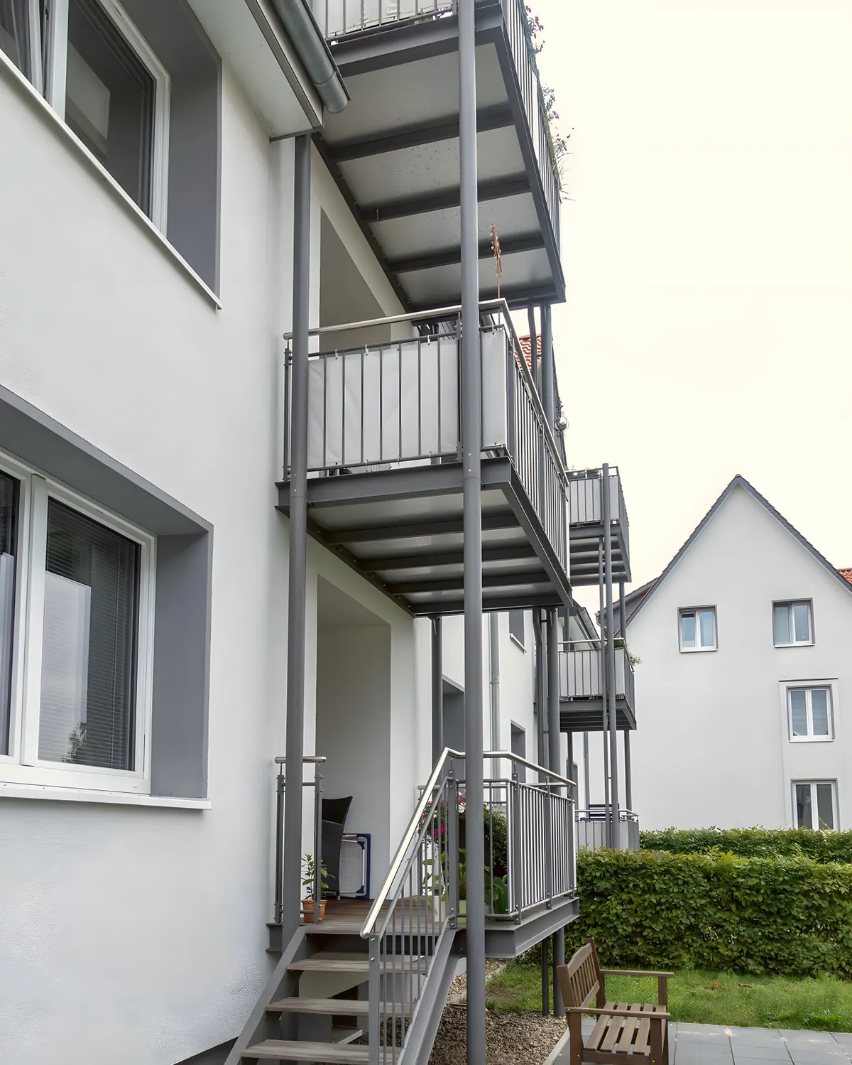 Übereinanderliegende Balkone in Anthrazit mit Stabgeländer an einem weißen Mehrfamilienhaus, unten eine Außentreppe in den Garten. Gebaut in Lüneburg.