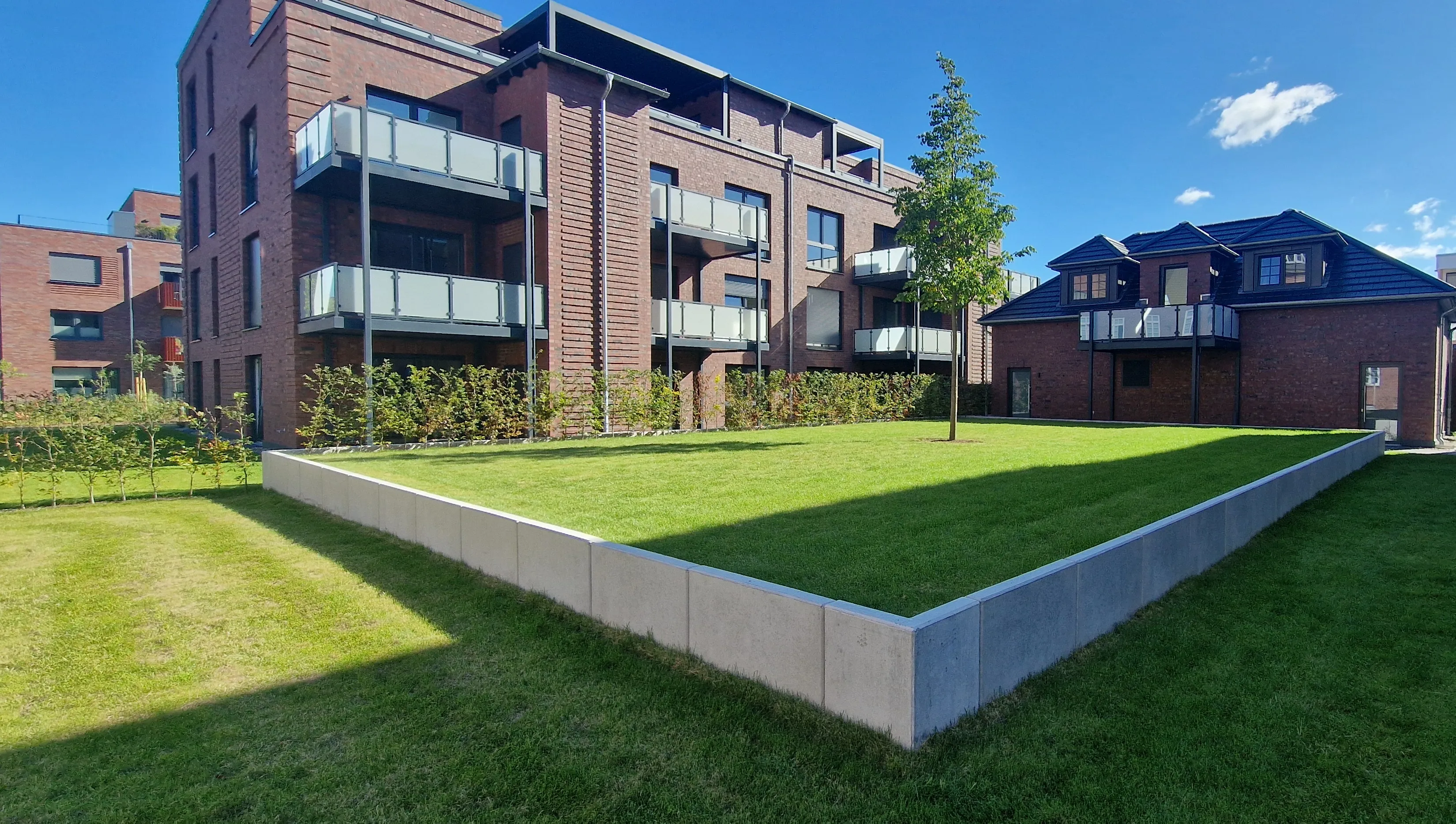 Wohnanlage aus Klinker mit mehreren übereinanderliegenden Vorstellbalkonen und Glasgeländern, davor ein neu angelegter Innenhof mit Rasen.